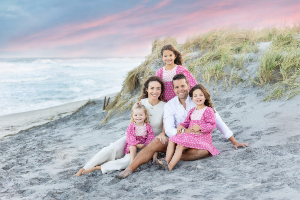 Family of five sitting by dunes on beach in pink and white colors
